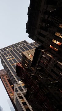 Two tall buildings are visible in the city during the late afternoon. One building has visible fire escapes. The sky is clear and blue.