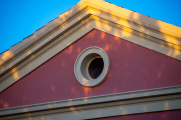 Architectural detail shows a red building facade with a round window, cornice, and blue sky backdrop. © AgioiDouloi