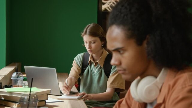 Rack focus shot of Caucasian female teenager writing in notebook and typing on laptop with Black student in foreground. Ideal for educational marketing, e-learning platforms, and university promos