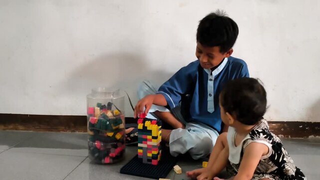 A young schoolboy wearing a blue uniform sits indoors with colorful building blocks while another child is nearby, capturing a candid childhood play moment.