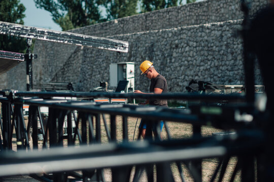Event crew member wearing hard hat working on stage setup, preparing outdoor concert rigging and sound equipment
