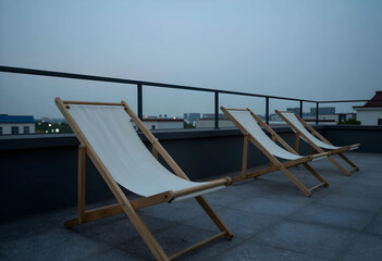 Fototapeta premium Three empty wooden deck chairs with white canvas on a rooftop terrace at dusk, urban skyline in the background, the urban skyline with buildings and a dark sky forms the background