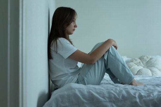 Depressed teenage girl sitting alone on a bed at home, feeling sad and overwhelmed, mental health concept