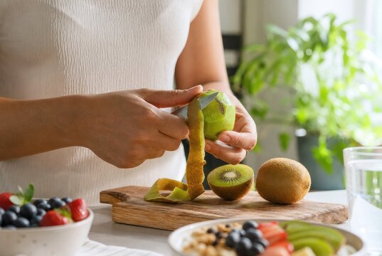 Fresh kiwi fruit being peeled by hands on wooden cutting board surrounded by healthy fruits and nuts in bright modern kitchen with indoor plants
