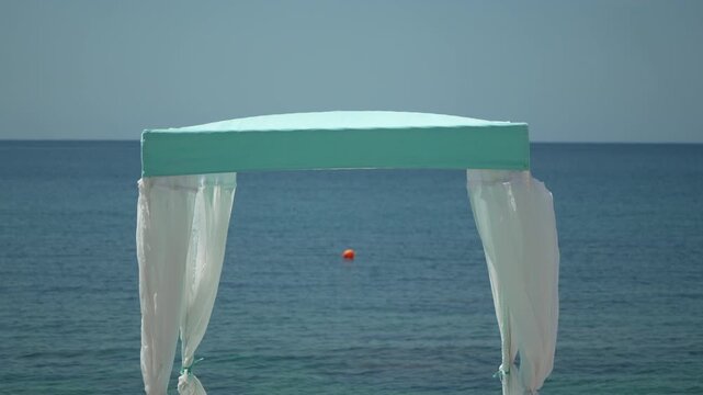Mediterranean summer breeze moves sheer curtains of a beach pavilion overlooking the calm sea. Static eye-level shot.