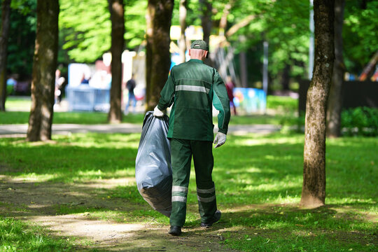Municipal park sanitation worker in green uniform carrying large black plastic trash bag along dirt path in park. Janitor collecting trash, carrying heavy garbage bag. Park maintenance service worker