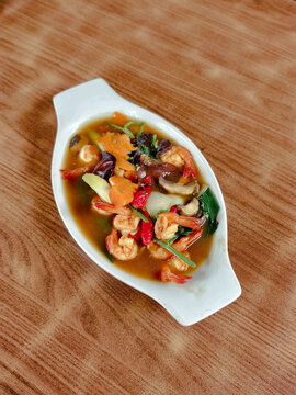Top view of delicious Capcay or stir-fried mixed vegetables with fresh prawns, carrots, and mushrooms served on a white ceramic plate over a wooden background. Healthy Asian seafood cuisine.