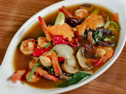 Top view of delicious Capcay or stir-fried mixed vegetables with fresh prawns, carrots, and mushrooms served on a white ceramic plate over a wooden background. Healthy Asian seafood cuisine.