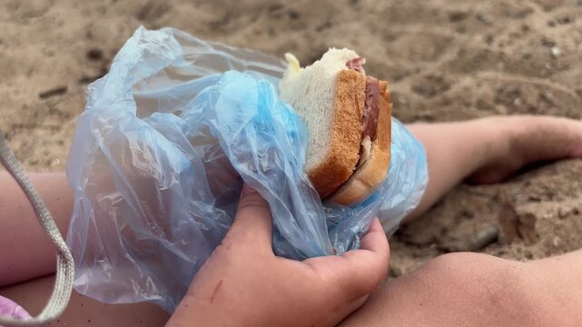 Child holding a sausage sandwich wrapped in a plastic bag on a sandy beach, casual snack, family outing, summer food.