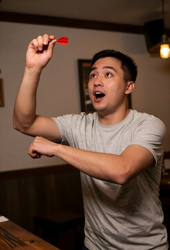 Excited young asian man playing darts with an open mouth expression, focused on the game in a casual setting