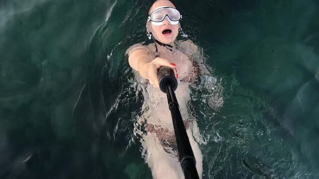 Underwater selfie perspective of woman in goggles and bikini holding monopod while swimming in dark sea. High angle action camera shot.