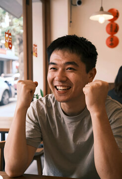 Excited young asian man with clenched fists celebrating success and achievement in a casual indoor setting, He expresses joy and enthusiasm in a casual indoor setting, possibly a cafe