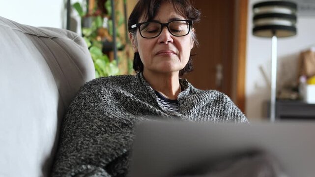 Focused senior businesswoman with glasses using a laptop computer while sitting comfortably on a couch, telecommuting and managing her business tasks with a serious expression from her home