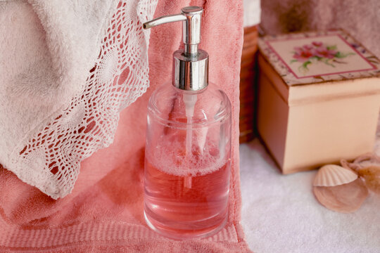Pink liquid soap dispenser standing on soft towels with a decorative lace edge and seashell, creating a serene atmosphere for beauty and self-care rituals