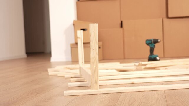 Wooden floor fills the room. Planks and a wooden frame structure scatter on the floor. Green drill sits nearby. Cardboard boxes stack against the wall