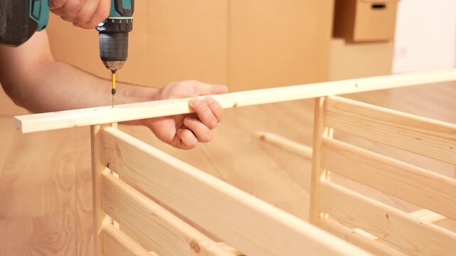 Person holds a wooden plank steady. Drill bit drops wood shavings. Wooden cabinet with drawers fills the background. This shows construction work in progress. Ideal for DIY home repair projects