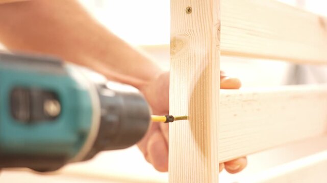 Close-up shot of hands drilling into a wooden plank. Drill bit secures the vertical wood piece. Hands guide the structure during assembly. Ideal for home improvement or DIY projects