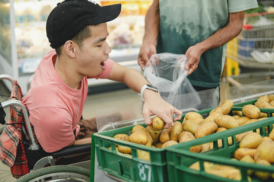 Happy young Asian man in a wheelchair choosing fresh potatoes at the supermarket, shopping for organic groceries with an assistant, concept of inclusive lifestyle and accessibility.