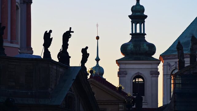 Silhouetted baroque statues on a church gable with a green onion-domed bell tower behind at dawn. Zoom out effect. Prague, Czech Republic