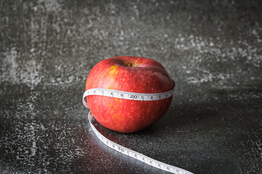 Fresh red apple wrapped in measuring tape on a dark gray studio background conveys mindful nutrition, portion control and a calm commitment to wellness