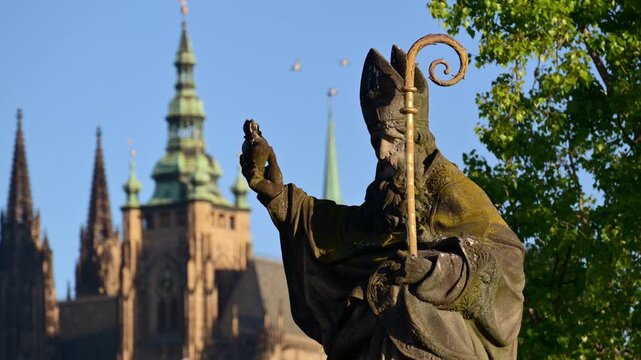 A weathered baroque bishop statue with a gilded crozier on Charles Bridge, St. Vitus Cathedral behind, Prague, Czech Republic