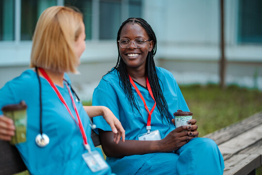 Two diverse female nurses smiling, communicating, and relaxing during a coffee break outdoors
