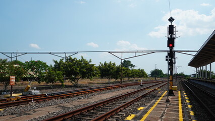 Red railway signal light stands along train station tracks during bright daytime. Eye level wide shot reveals multiple steel rails, platform yellow safety line, and green trees under clear blue sky. © wahid HA