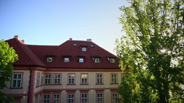 A baroque building with a red tiled roof and dormer windows beside a sunlit green tree, Prague, Czech Republic
