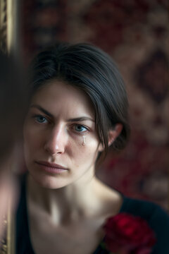 Single tear running down cheek of a sad woman looking at her reflection in a mirror with a red rose, emotional portrait of grief, melancholy, heartbreak and sorrow, intimate mood close-up photograph