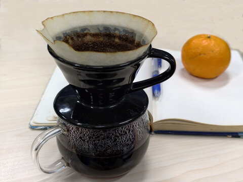 Pour over coffee brewer dripping into glass mug on work desk with notebook and orange fruit