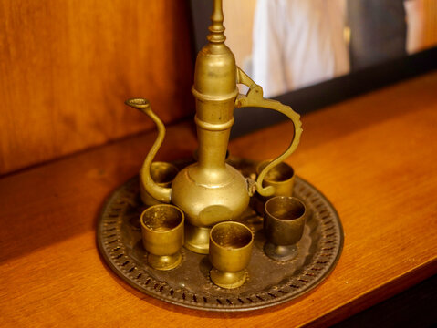 A high-angle, close-up photograph of an antique brass coffee or tea set, featuring a tall, elegant dallah-style pot surrounded by small goblets on an ornate, patterned serving tray against a wooden ba