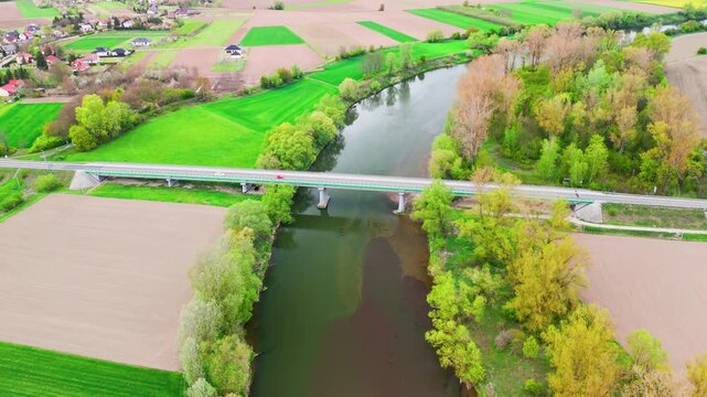 Aerial view of river with bridge crossing through farmland and colorful autumn trees near small village