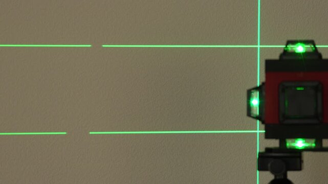 Red device with green lights rests on a beige surface. Horizontal green grid lines cross the scene. It has a square screen and black parts. This tool appears to measure distance accurately