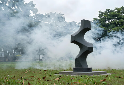 Dark gray stone sculpture with geometric shapes in a park with Smoke and trees, Smoke drifts around the artwork and trees in the background