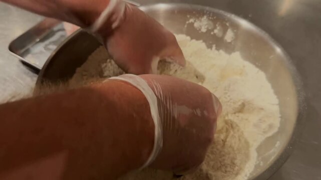 A culinary worker wearing sanitary protective gloves handles poultry portions through a multi-step station involving dry flour coating, liquid egg wash, and textured breadcrumb layers