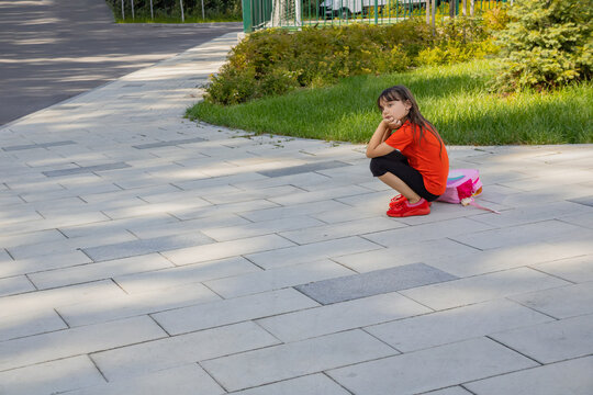 Sad little girl with pink backpack squatting on paved sidewalk outdoors, thoughtful bored child waiting and back to school concept