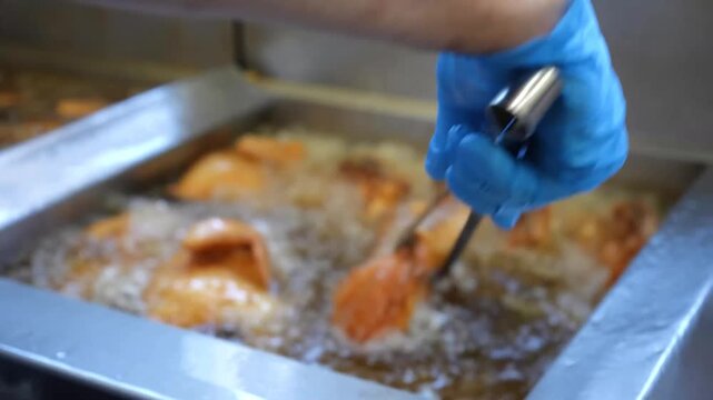 Raw chicken pieces are submerged into hot boiling oil vats using metal tongs inside a commercial restaurant kitchen environment for professional frying