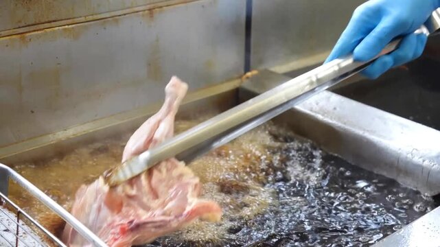 Raw chicken pieces are submerged into hot boiling oil vats using metal tongs inside a commercial restaurant kitchen environment for professional frying