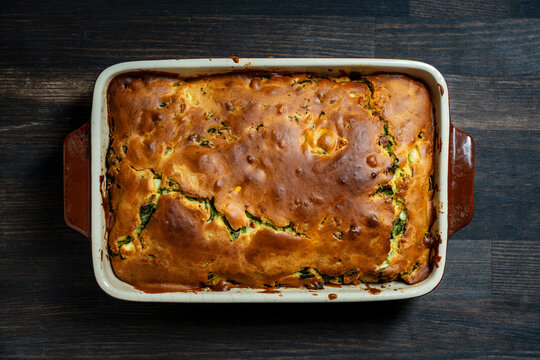 Homemade savory pie with golden crust in baking dish. Top view of freshly baked golden brown savory pie with green vegetables and egg filling in ceramic pan placed on rustic wooden table, close up