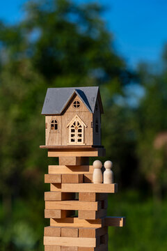 A toy wooden house model and peg dolls on top of block tower outdoors with blue sky and green trees background representing real estate risk, closeup