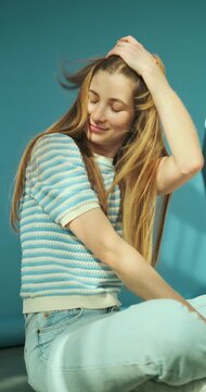 Vertical video of young happy blonde woman with long healthy hair in striped top and jeans sitting on the floor in studio on blue background