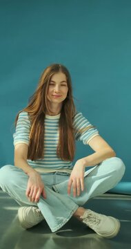 Vertical video of young happy blonde woman with long healthy hair in striped top and jeans sitting on the floor in studio on blue background