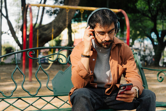 Young man with headphones using cell phone sitting on park bench