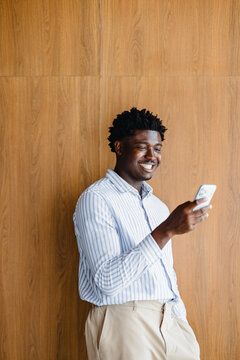 Smiling Man Looking at His Phone While Standing by a Wooden Wall