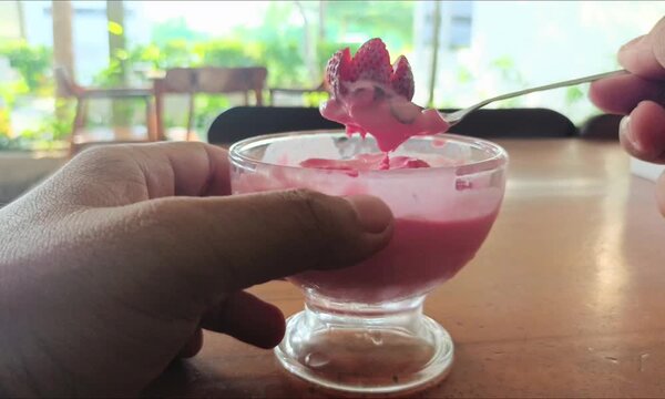 Person scooping strawberry ice cream from glass bowl on wooden table