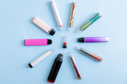 A flat-lay view of multiple disposable electronic cigarettes and one vape pod arranged in a circle on a light blue background. High-key product photography.