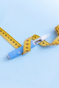 Weight loss injection pen wrapped in a yellow measuring tape on blue background. Concept of health risk, dangerous side effects, medical complications, drug misuse, and extreme slimming methods.