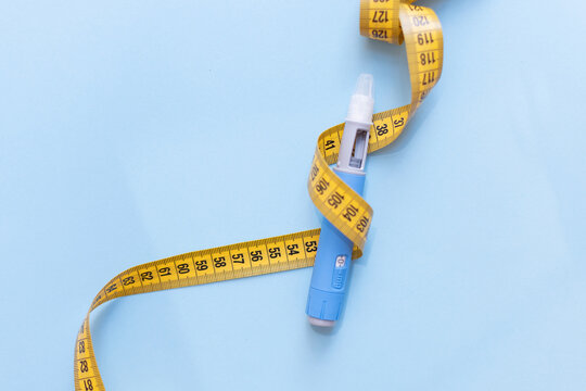 Blue injection pen wrapped in a yellow measuring tape on light blue background. Concept of health risk, dangerous side effects, medical complications, drug abuse, and extreme slimming methods.
