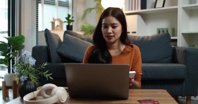 Young asian woman working remotely on laptop and phone in modern home living room, freelancer productivity