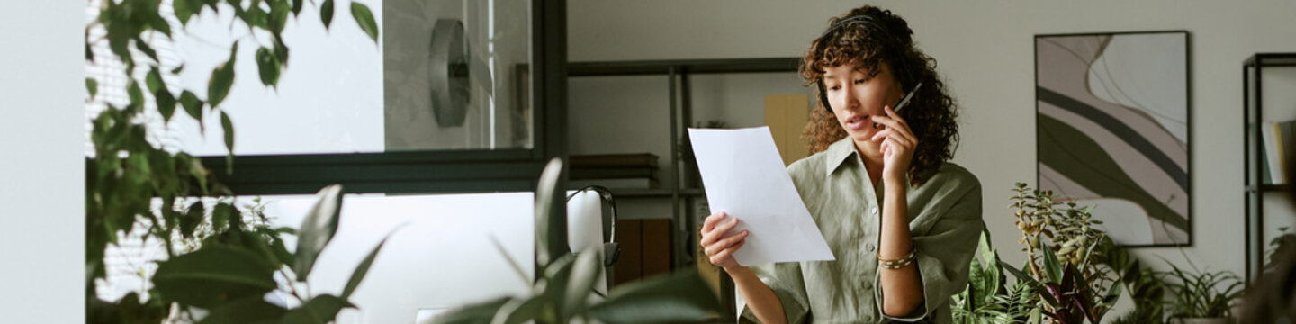 Header of Biracial young adult woman working in call center wearing headset and reading document. Image suitable for customer service training, and telemarketing campaigns